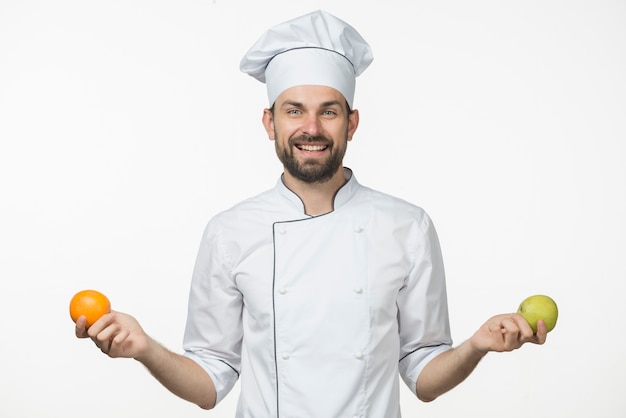 Бесплатное фото Улыбаясь молодой мужской шеф-повар холдинг и спелые оранжевые и зеленое яблоко в руке