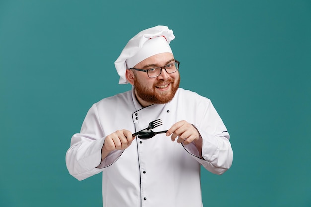 Smiling young male chef wearing glasses uniform and cap holding fork and spoon looking at camera rubbing them against each other isolated on blue background