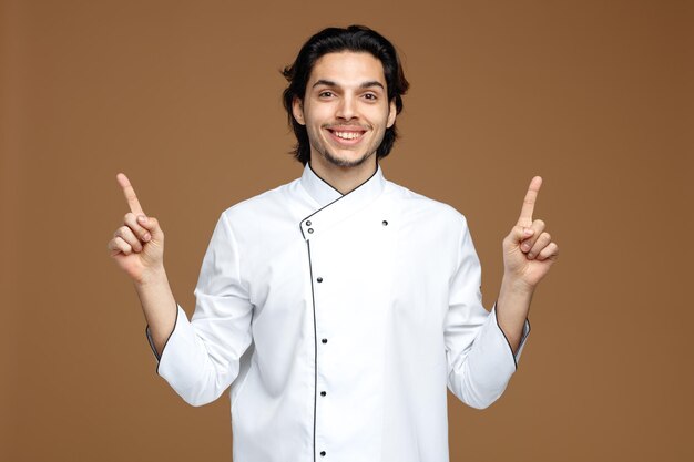 갈색 배경에 고립 된 손가락을 가리키는 카메라를보고 유니폼을 입고 웃는 젊은 남성 요리사