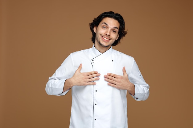 smiling young male chef wearing uniform looking at camera pointing at himself with hands isolated on brown background