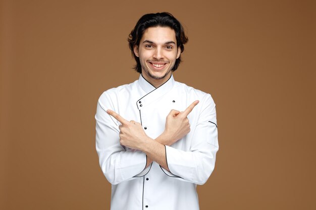 갈색 배경에 고립 된 측면을 가리키는 카메라를보고 유니폼을 입고 웃는 젊은 남성 요리사