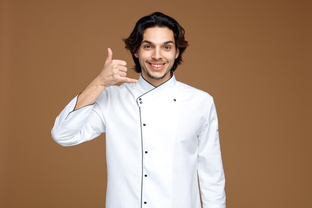 smiling young male chef wearing uniform looking at camera showing call gesture isolated on brown background