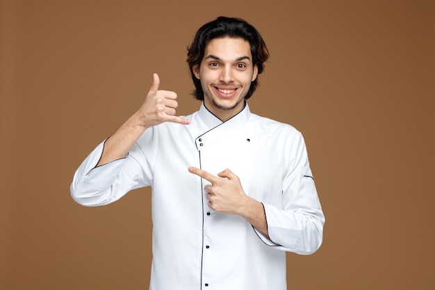 갈색 배경에 고립 된 쪽을 가리키는 호출 제스처를 보여주는 카메라를보고 유니폼을 입고 웃는 젊은 남성 요리사