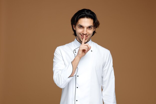 smiling young male chef wearing uniform looking at camera showing silence gesture isolated on brown background