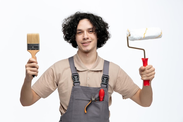 Smiling young male construction worker wearing uniform holding paint brush and paint roller looking at camera with screwdriver and pliers in pocket isolated on white background