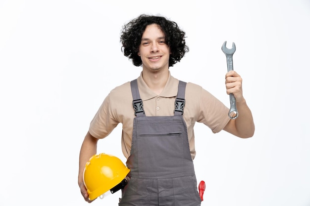 Smiling young male construction worker wearing uniform holding safety helmet showing spanner to camera looking at camera with screwdriver in pocket isolated on white background