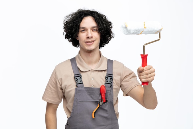 Smiling young male construction worker wearing uniform looking at camera showing paint roller to camera with screwdriver and pliers in pocket isolated on white background
