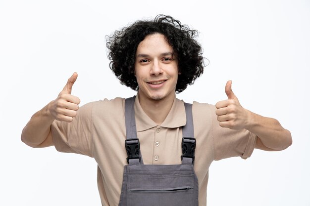 Улыбающийся молодой мужчина-строитель в униформе смотрит в камеру, показывая большие пальцы на белом фоне