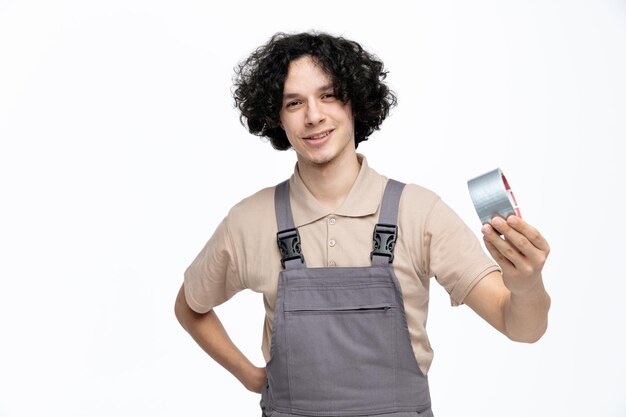 Smiling young male construction worker wearing uniform stretching scotch tape towards camera looking at camera while keeping hand on waist isolated on white background