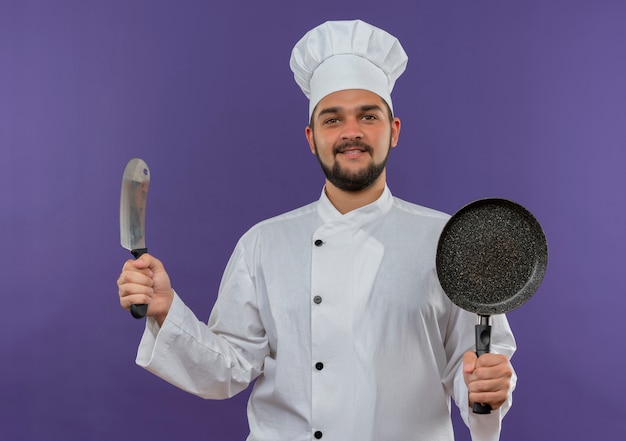 칼과 보라색 공간에 고립 된 프라이팬을 들고 요리사 유니폼에 웃는 젊은 남성 요리사