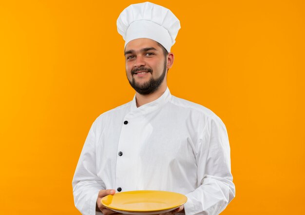 Smiling young male cook in chef uniform holding empty plate isolated on orange space