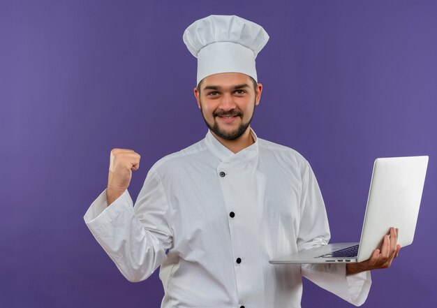 ラップトップを保持し、紫色の空間で隔離の拳を握り締めるシェフの制服を着た若い男性料理人の笑顔