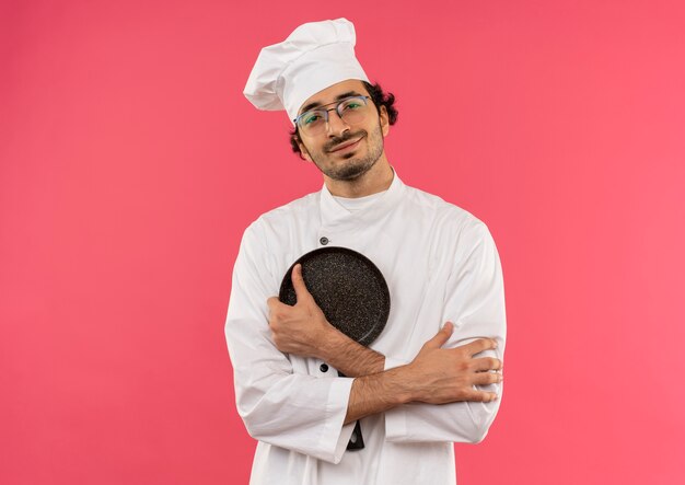 smiling young male cook wearing chef uniform and glasses crossing hands and holding frying pan