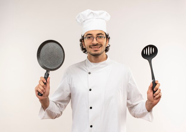 Smiling young male cook wearing chef uniform and glasses holding frying pan and spatula isolated on white wall