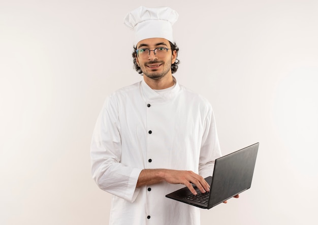 Sorridente giovane cuoco maschio indossando l'uniforme del cuoco unico e vetri che tengono laptop isolato sulla parete bianca