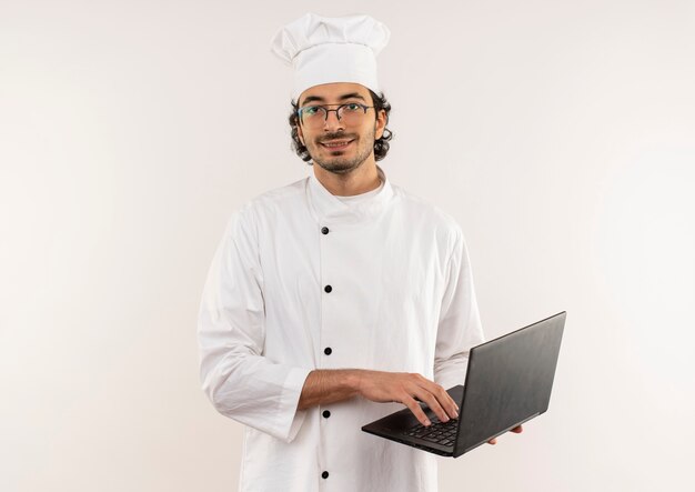 Smiling young male cook wearing chef uniform and glasses holding laptop isolated on white wall