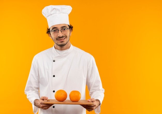 Smiling young male cook wearing chef uniform and glasses holding orange on cutting board isolated on yellow wall