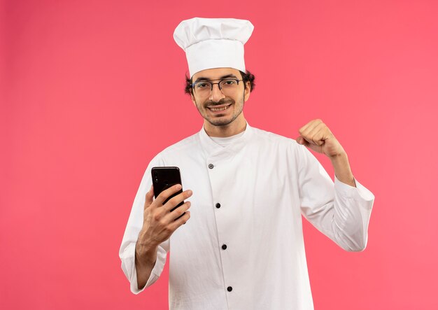 Улыбающийся молодой мужчина-повар в униформе шеф-повара и очках держит телефон и делает сильный жест, изолированный на розовой стене