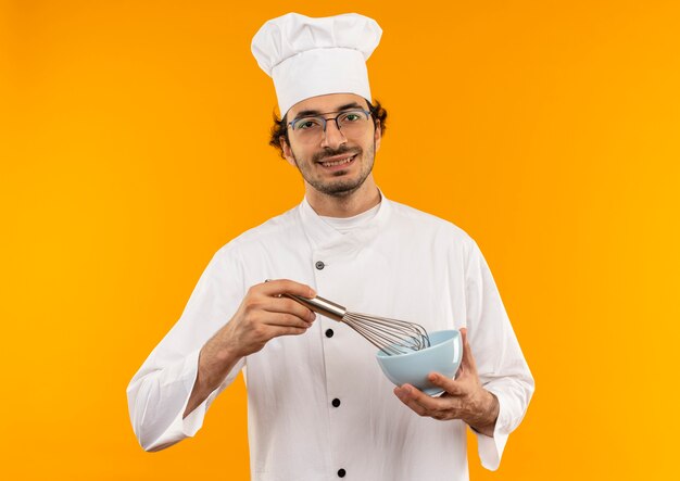 Улыбающийся молодой мужчина-повар в униформе шеф-повара и в очках держит венчик и миску, изолированные на желтой стене