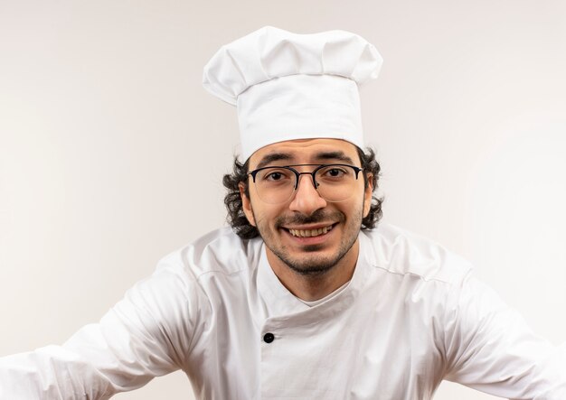Smiling young male cook wearing chef uniform and glasses isolated on white wall