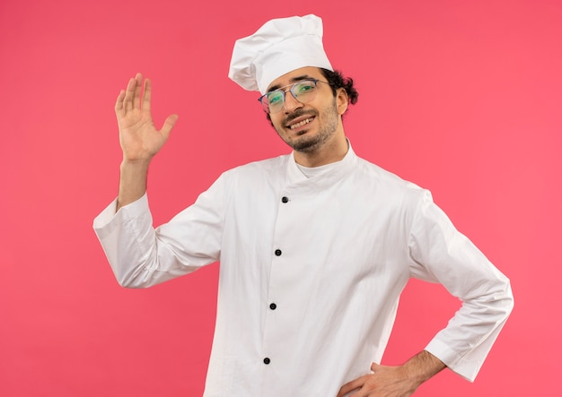 Smiling young male cook wearing chef uniform and glasses raising hand and putting another hand on hip on pink