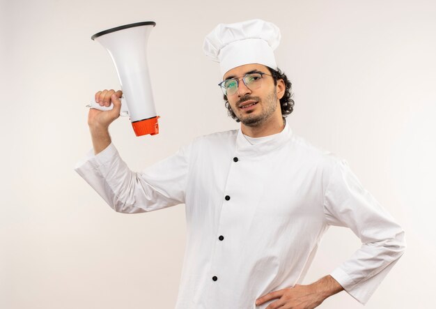 Smiling young male cook wearing chef uniform and glasses raising loudspeaker putting hand on hip isolated on white wall