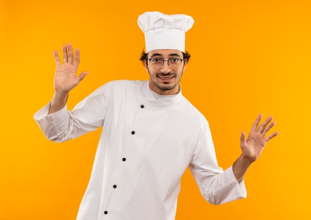 Smiling young male cook wearing chef uniform and glasses spreads hands isolated on yellow wall