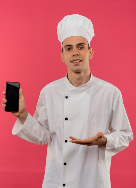 Smiling young male cook wearing chef uniform holding and points with hand to phone