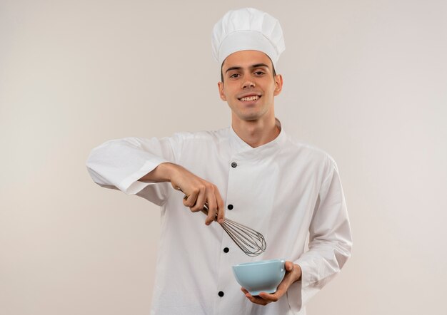 Smiling young male cook wearing chef uniform holding whisk with bowl
