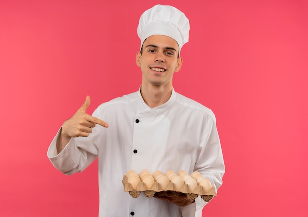 Smiling young male cook wearing chef uniform points finger to batch of eggs in his hand with copy space