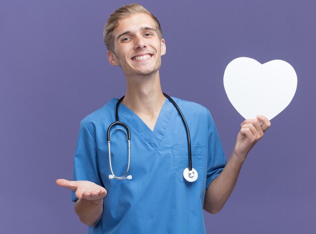 Smiling young male doctor wearing doctor uniform with stethoscope holding heart shape box points with hand at camera isolated on blue wall
