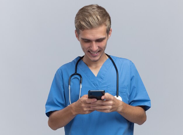 Smiling young male doctor wearing doctor uniform with stethoscope holding and looking at phone isolated on white wall with copy space