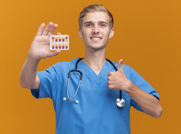Smiling young male doctor wearing doctor uniform with stethoscope holding pills showing thumb up isolated on orange wall