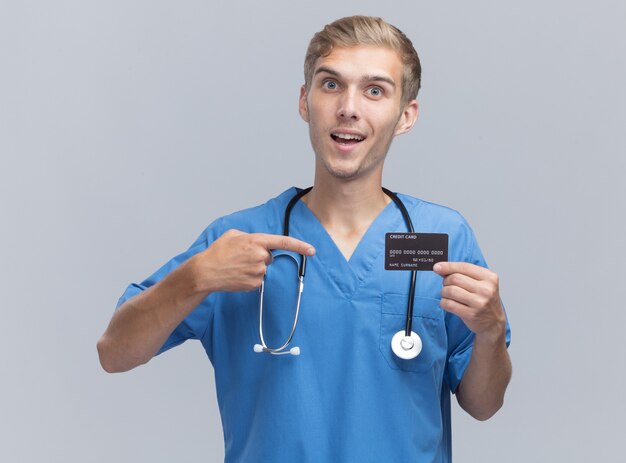 Улыбающийся молодой мужчина-врач в униформе врача со стетоскопом и указывает на кредитную карту, изолированную на белой стене