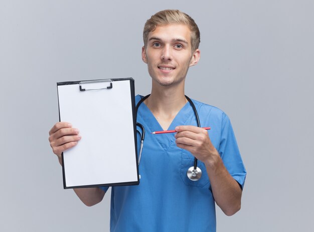 Sorridente giovane medico maschio indossando l'uniforme del medico con la tenuta dello stetoscopio e punti con la matita negli appunti isolato sulla parete bianca