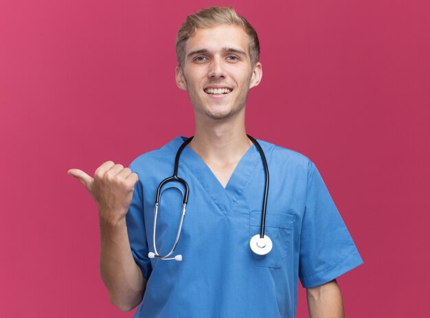 Smiling young male doctor wearing doctor uniform with stethoscope points at side isolated on pink wall with copy space