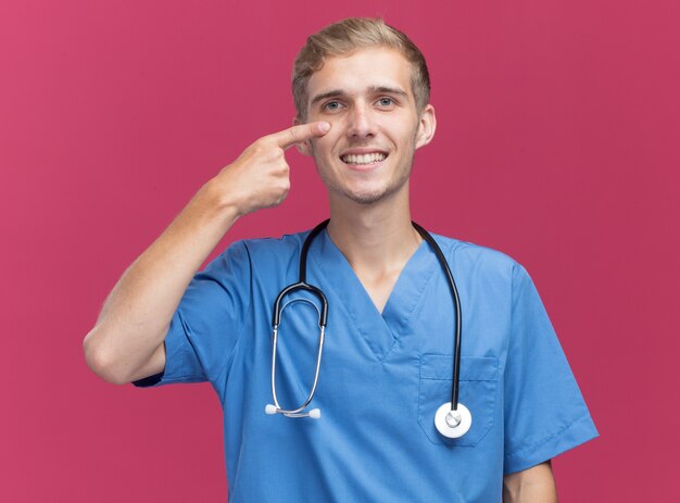 Smiling young male doctor wearing doctor uniform with stethoscope pulling down eye lid isolated on pink wall