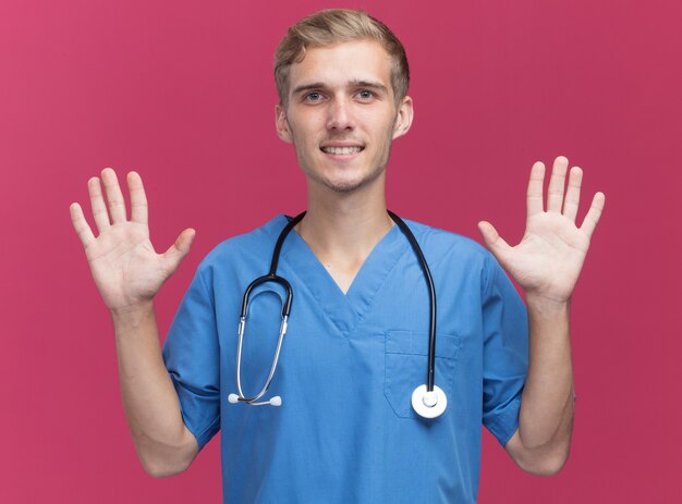 Smiling young male doctor wearing doctor uniform with stethoscope raising hands isolated on pink wall