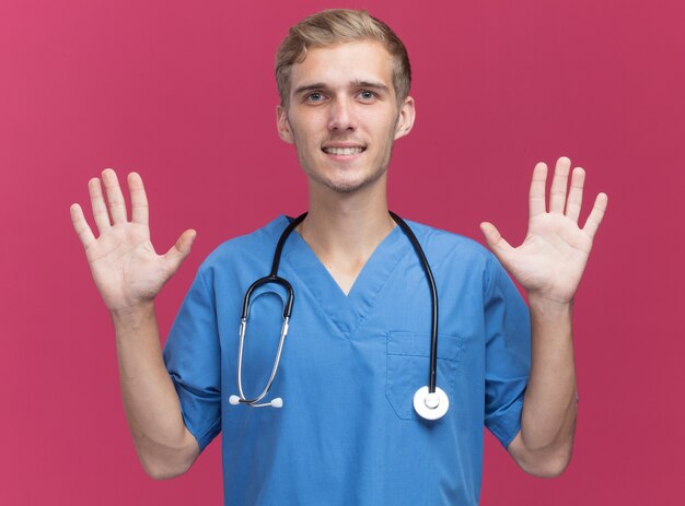 Улыбающийся молодой мужчина-врач в униформе врача со стетоскопом, поднимая руки, изолированные на розовой стене