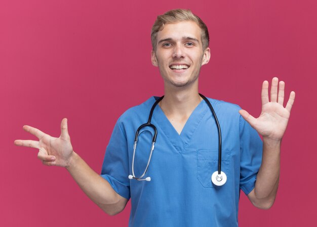 Smiling young male doctor wearing doctor uniform with stethoscope showing differents numbers isolated on pink wall