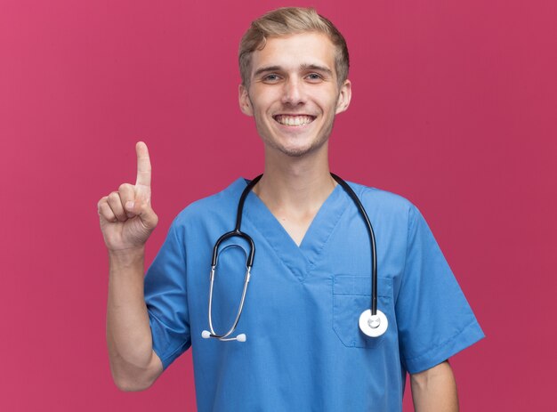 Smiling young male doctor wearing doctor uniform with stethoscope showing one isolated on pink wall
