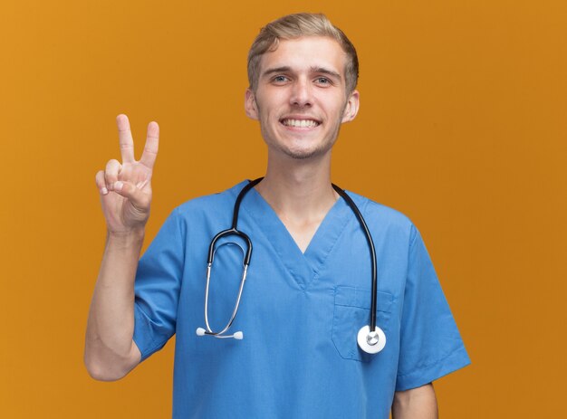 오렌지 벽에 고립 된 평화 제스처를 보여주는 청진 기와 의사 유니폼을 입고 웃는 젊은 남성 의사