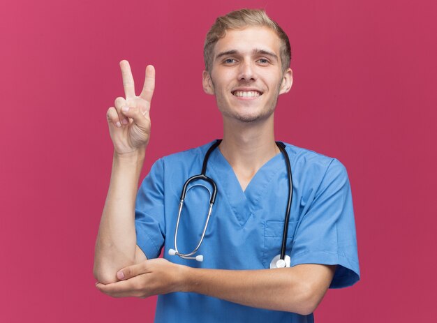 Smiling young male doctor wearing doctor uniform with stethoscope showing peace gesture isolated on pink wall