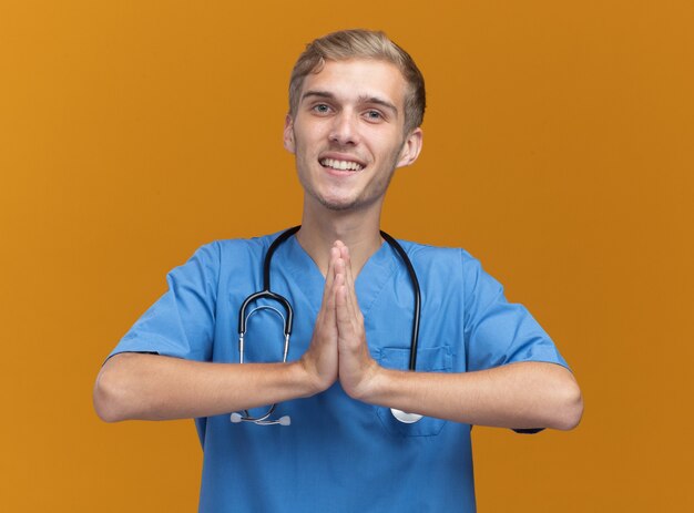オレンジ色の壁に分離された祈りのジェスチャーを示す聴診器と医師の制服を着て笑顔の若い男性医師