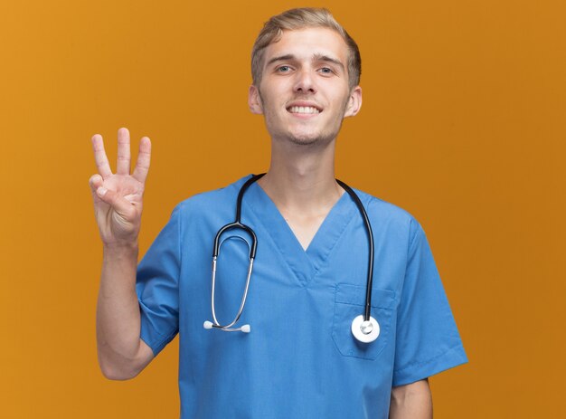 오렌지 벽에 고립 된 3을 보여주는 청진 기와 의사 유니폼을 입고 웃는 젊은 남성 의사