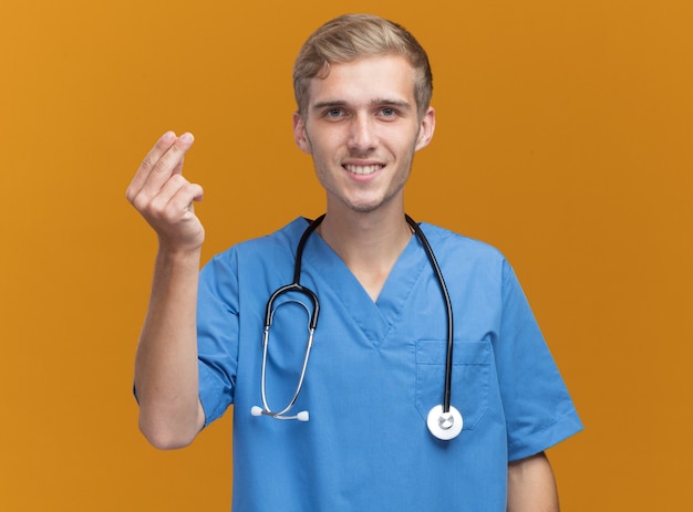 오렌지 벽에 고립 된 팁 제스처를 보여주는 청진기와 의사 유니폼을 입고 젊은 남성 의사 미소