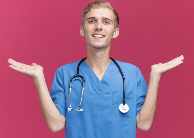 Smiling young male doctor wearing doctor uniform with stethoscope spreading hands isolated on pink wall