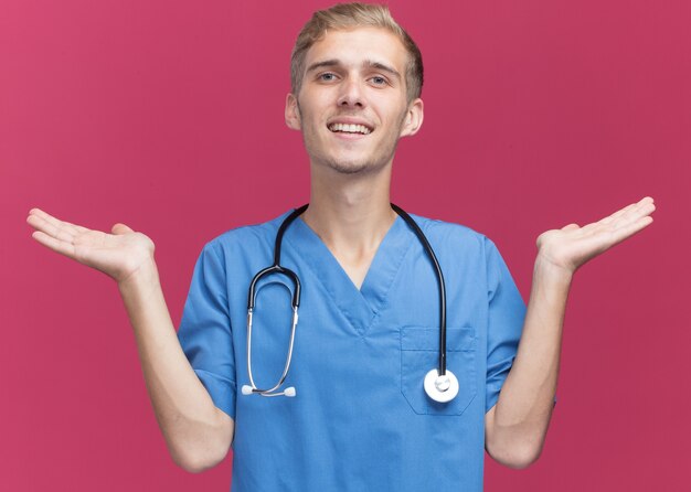 Улыбающийся молодой мужчина-врач в униформе врача со стетоскопом, разводя руками, изолированными на розовой стене