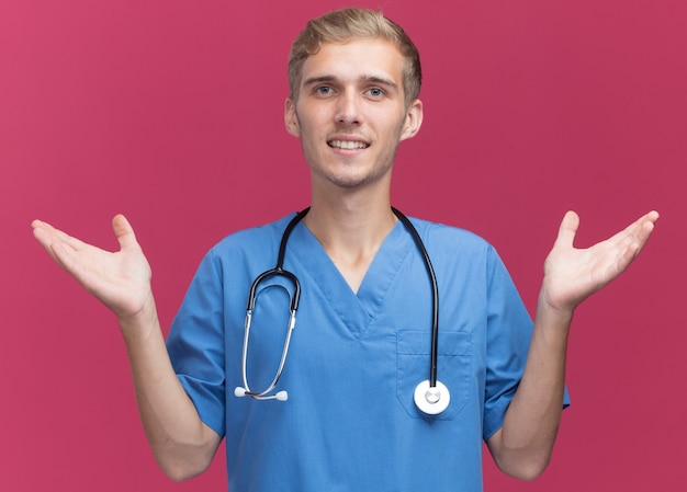 Smiling young male doctor wearing doctor uniform with stethoscope spreading hands isolated on pink wall