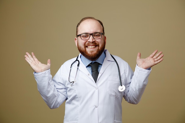 Smiling young male doctor wearing glasses lab coat and stethoscope around his neck looking at camera showing empty hands isolated on olive green background